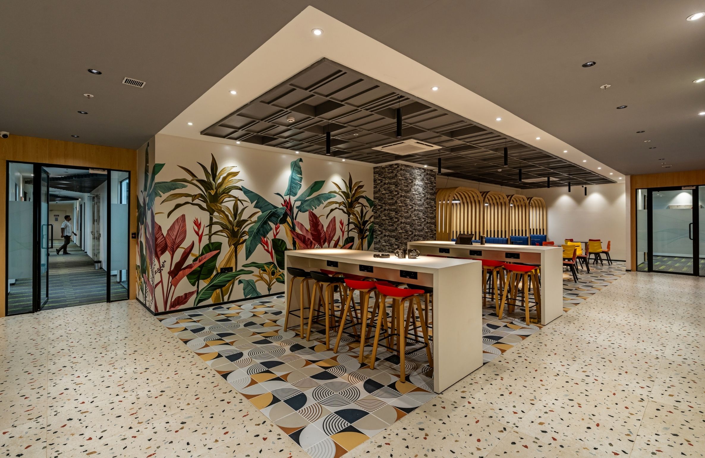 Modern office lounge and cafeteria area in an office space in Ameerpet, featuring tropical wall murals, high stools, patterned flooring, recessed ceiling lights, and a contemporary open seating layout.